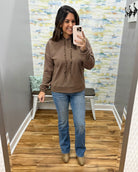 A woman takes a mirror selfie wearing a brown pullover hoodie with beaded drawstrings, mica bootcut jeans, and corkys tan ankle boots in a fitting room with wood floors and abstract wallpaper.