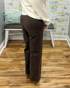 A person shown from the back wearing dark brown wide-leg pants, a cream knit sweater, and tan shoes, standing on a wood floor in an indoor room with a bench and patterned wall.