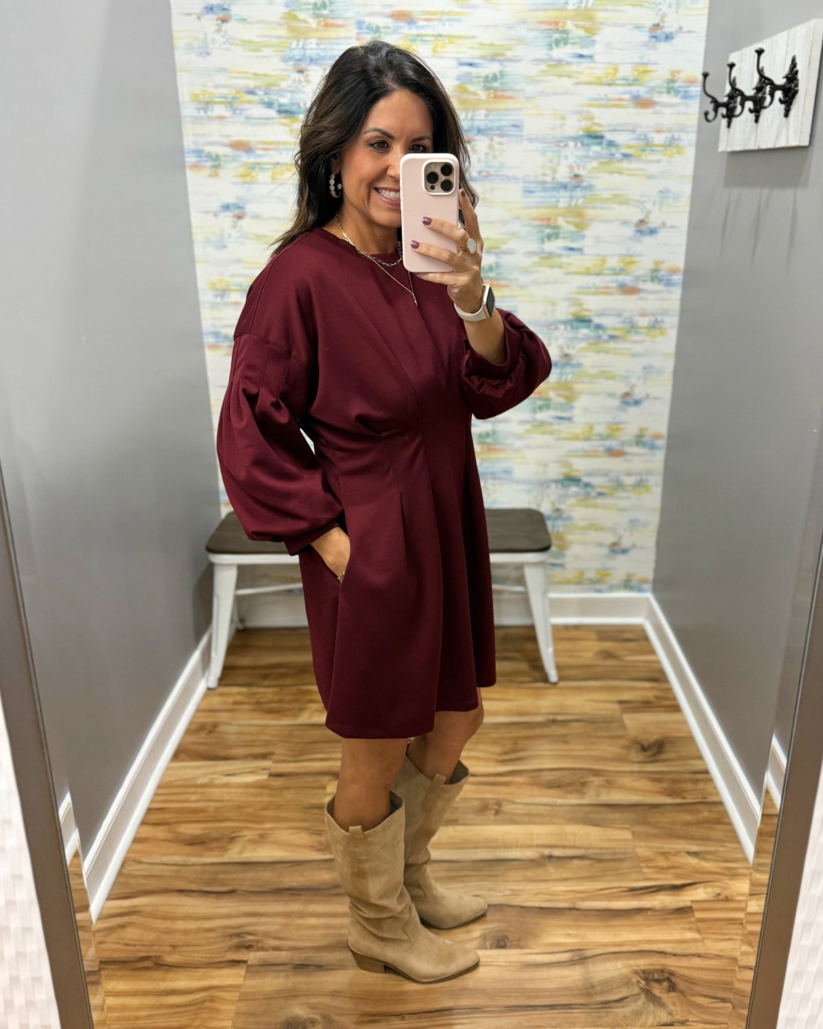 A woman takes a mirror selfie in a fitting room wearing a long-sleeve burgundy midi dress and beige knee-high boots.