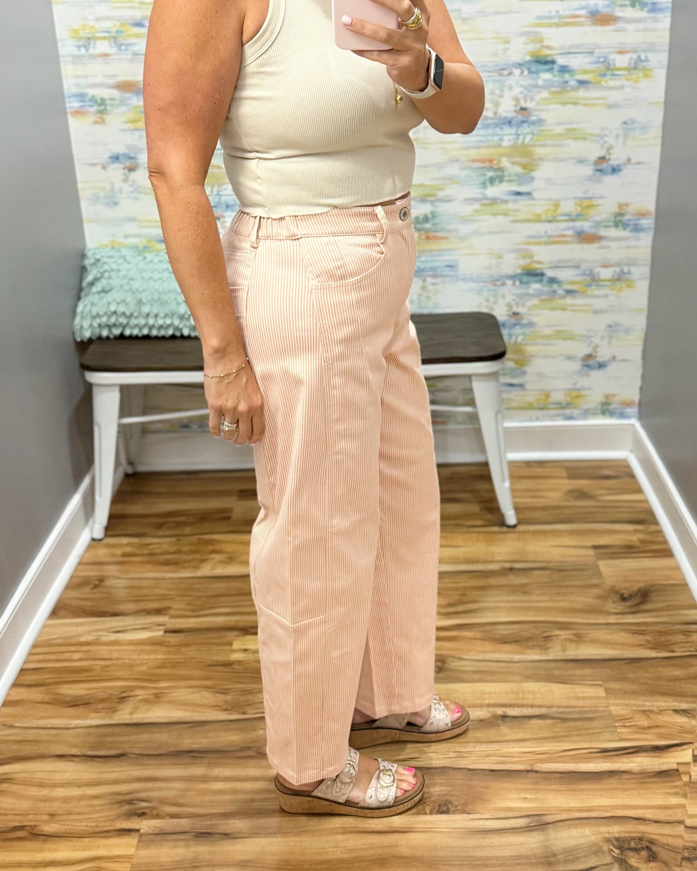 A woman modeling light peach corduroy wide-leg pants with a front slit, paired with a cream tank top and sandals, standing in a room with wood floors and a bench.