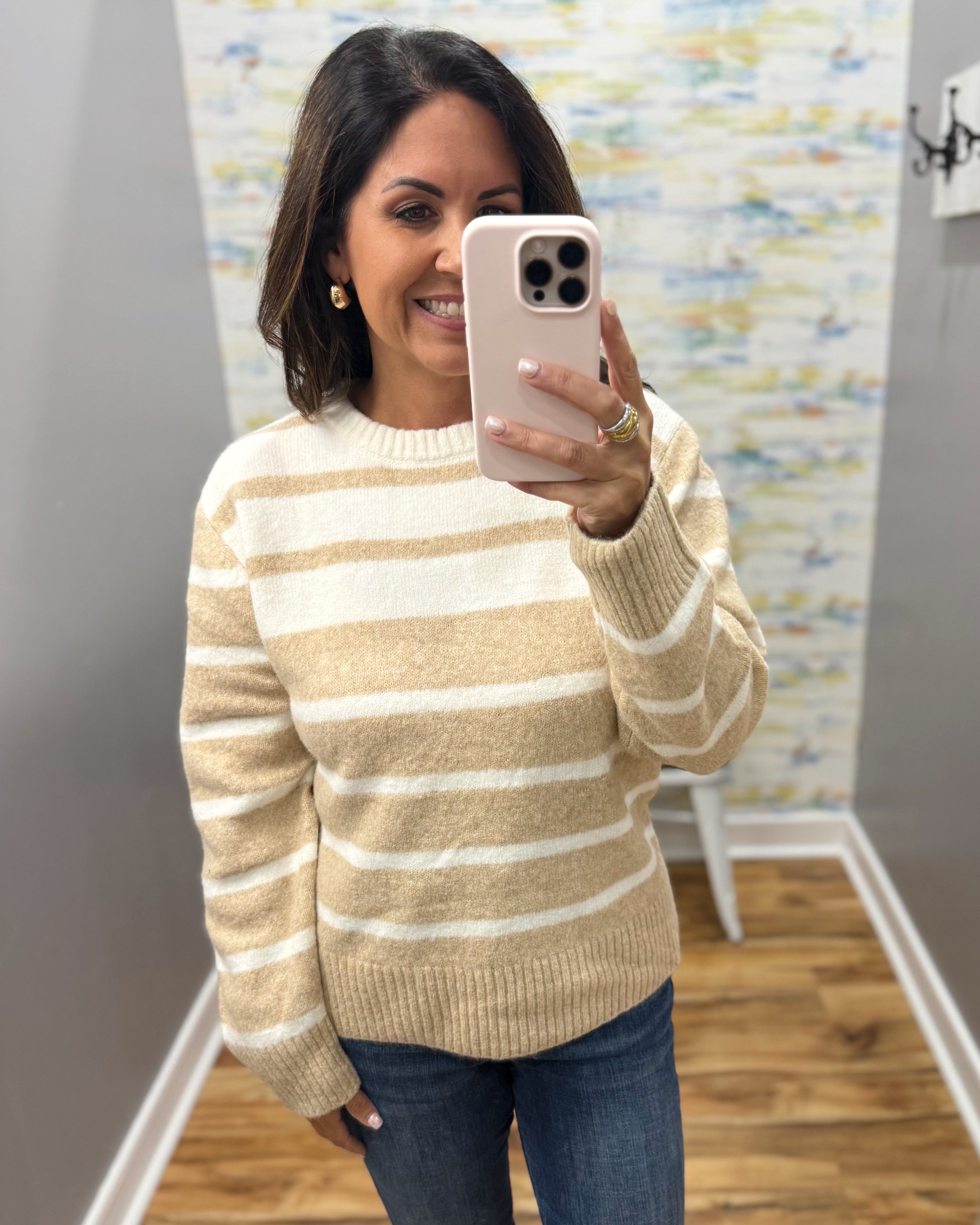 A woman takes a mirror selfie wearing a beige and white horizontally striped knit sweater, blue jeans, and a light pink phone case in a home setting.