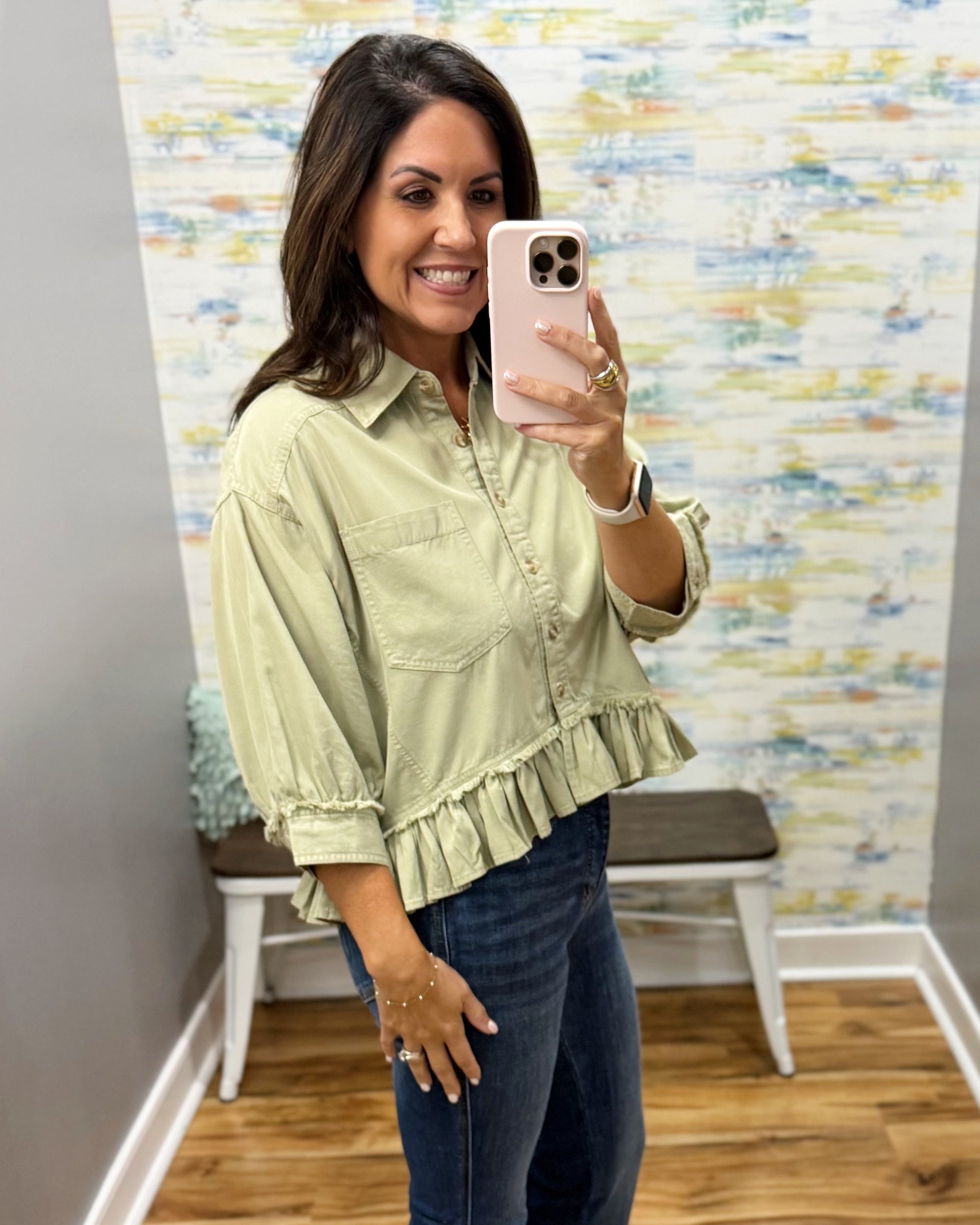 A woman takes a mirror selfie wearing a light sage green ruffled blouse and dark blue jeans in a home setting with patterned wallpaper and a wooden floor.