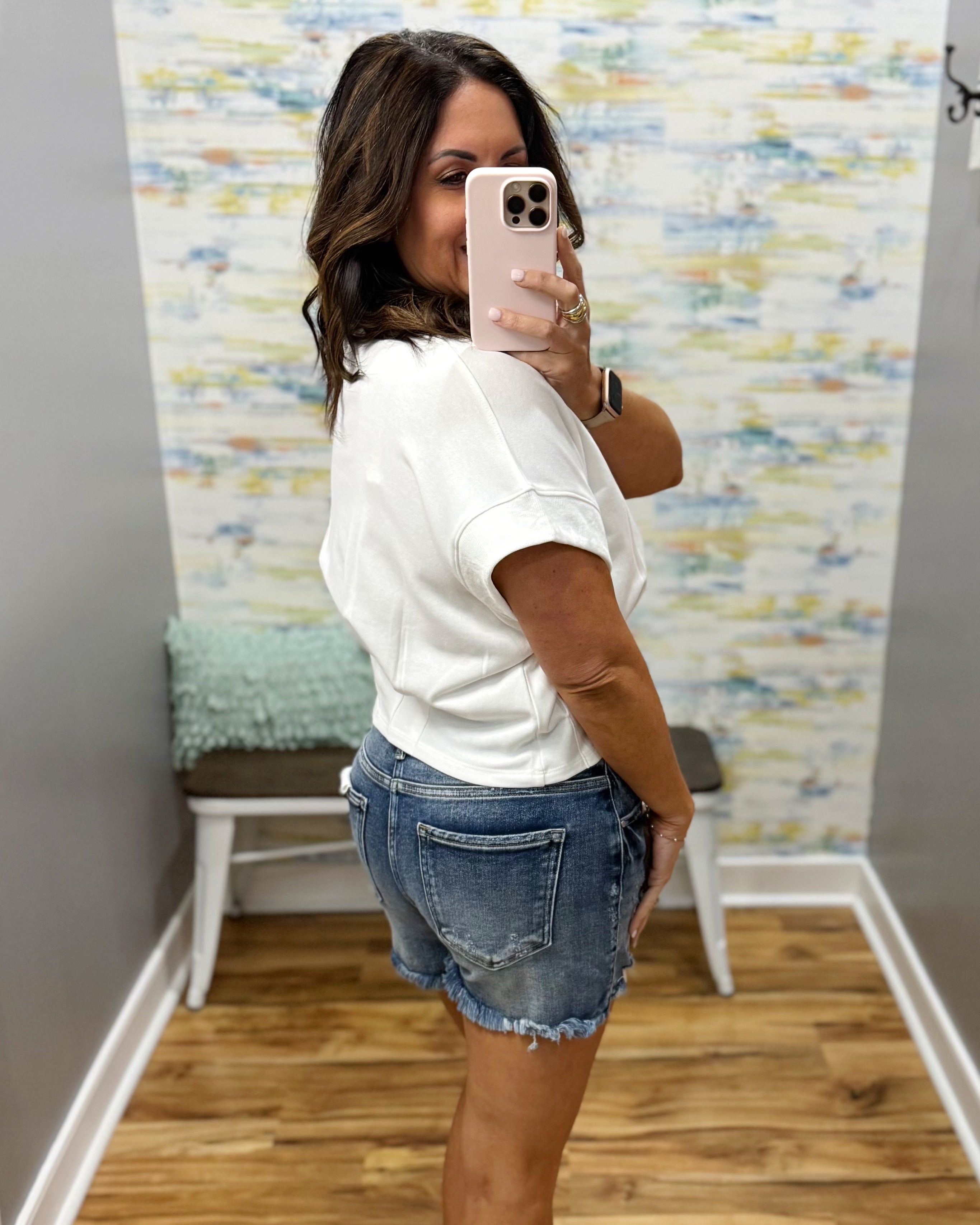 Woman taking a mirror selfie wearing a white t-shirt and denim shorts in a room with a colorful wall.