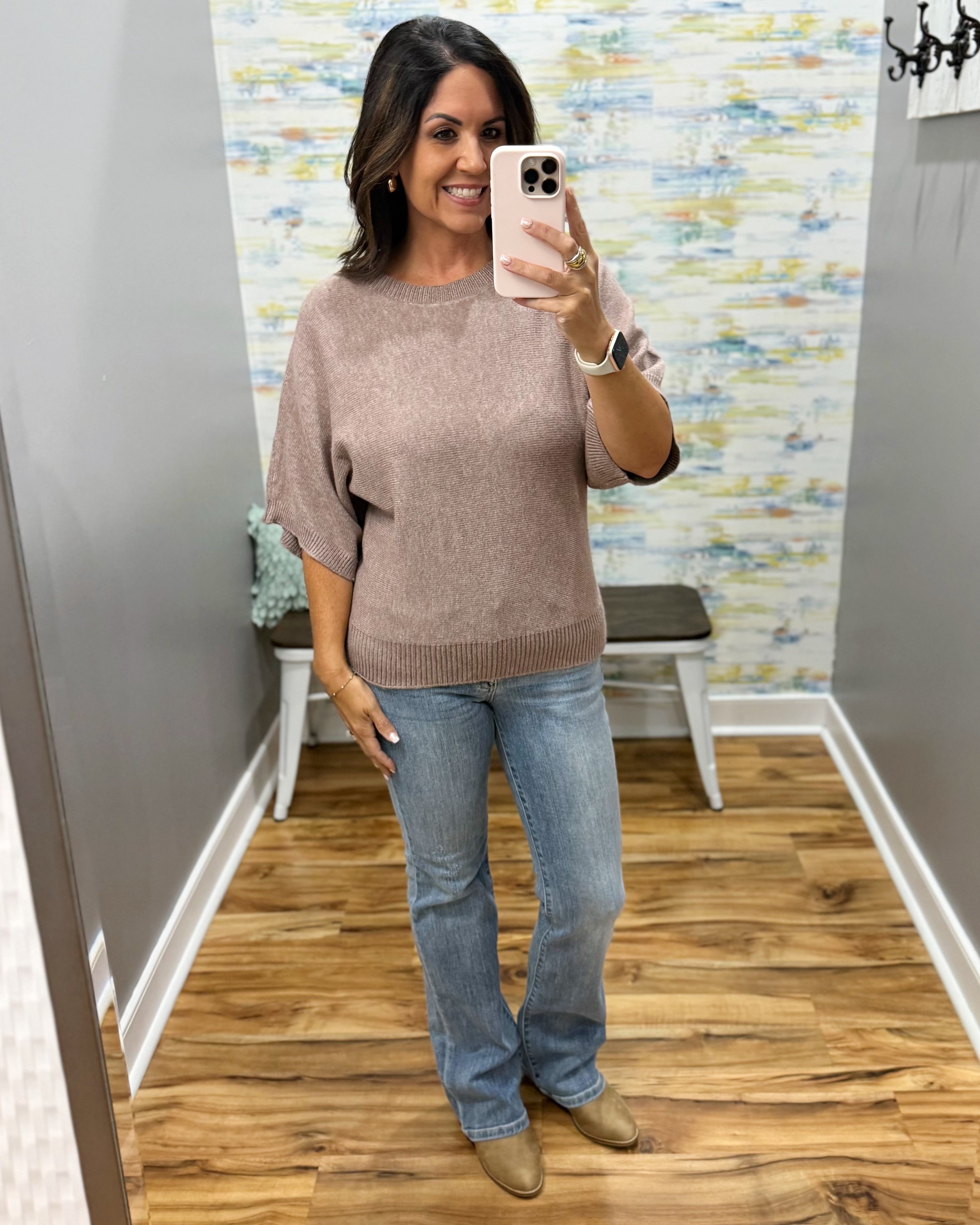 A woman takes a mirror selfie wearing a dusty rose knit sweater and light blue jeans in a home fitting room with wood floors and a patterned wallpaper background.