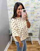 A woman takes a mirror selfie wearing a cream-colored knit sweater with dark brown polka dots and light blue jeans in a boutique setting.