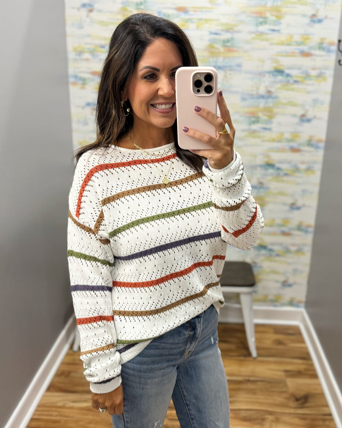 A woman takes a mirror selfie wearing a white knit sweater with horizontal stripes in orange, green, and navy blue, paired with blue jeans.