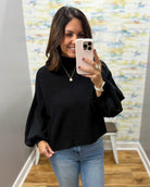 A woman takes a mirror selfie wearing a black long-sleeve mock neck top with dramatic balloon sleeves, paired with blue jeans. She is holding a light pink smartphone in front of her face.