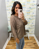 A woman takes a mirror selfie wearing a brown textured knit sweater and light blue jeans in a home setting with a patterned wallpaper background.