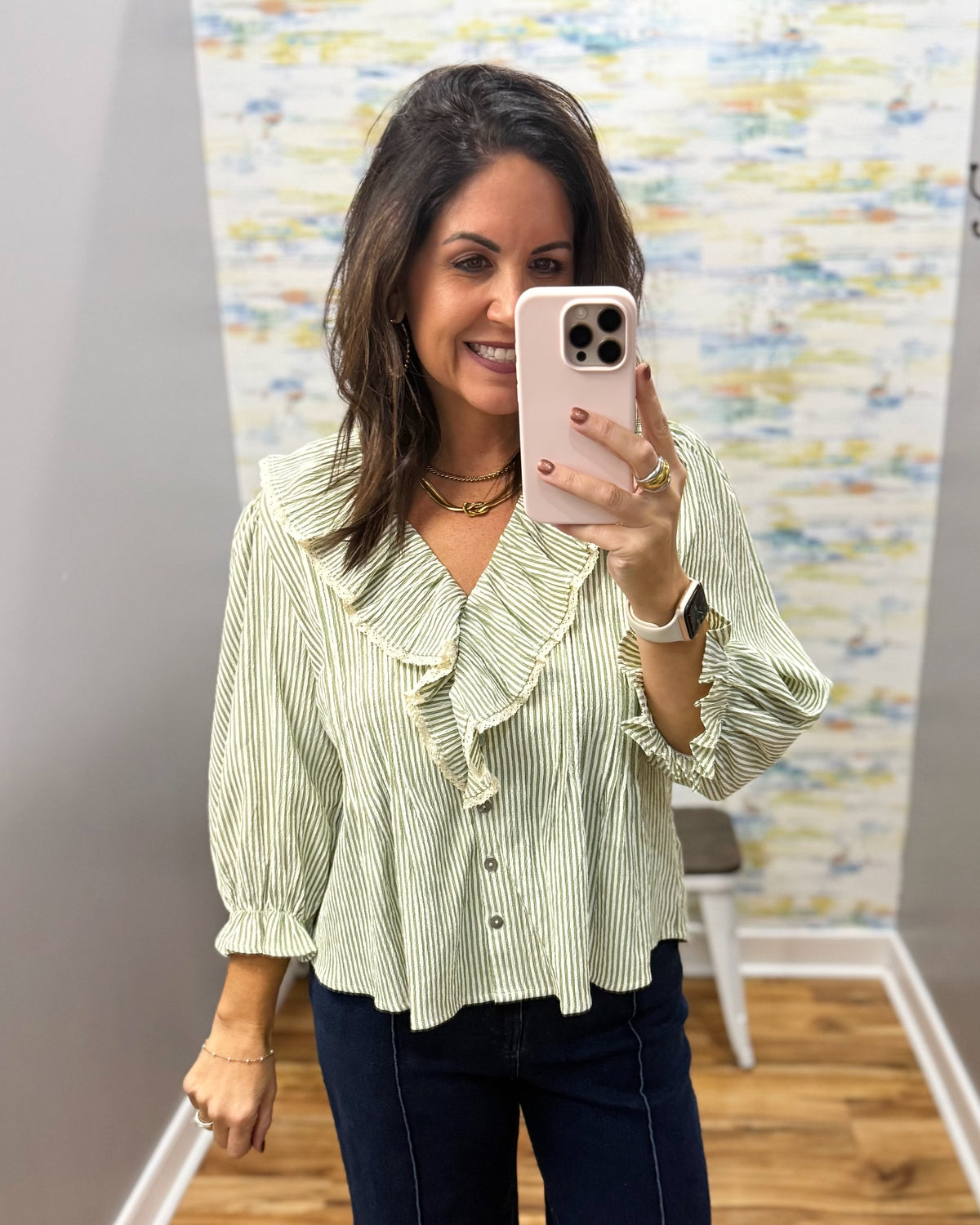 A woman takes a mirror selfie wearing a light sage green and white vertically striped blouse with ruffle details on the collar and cuffs, paired with dark blue jeans.