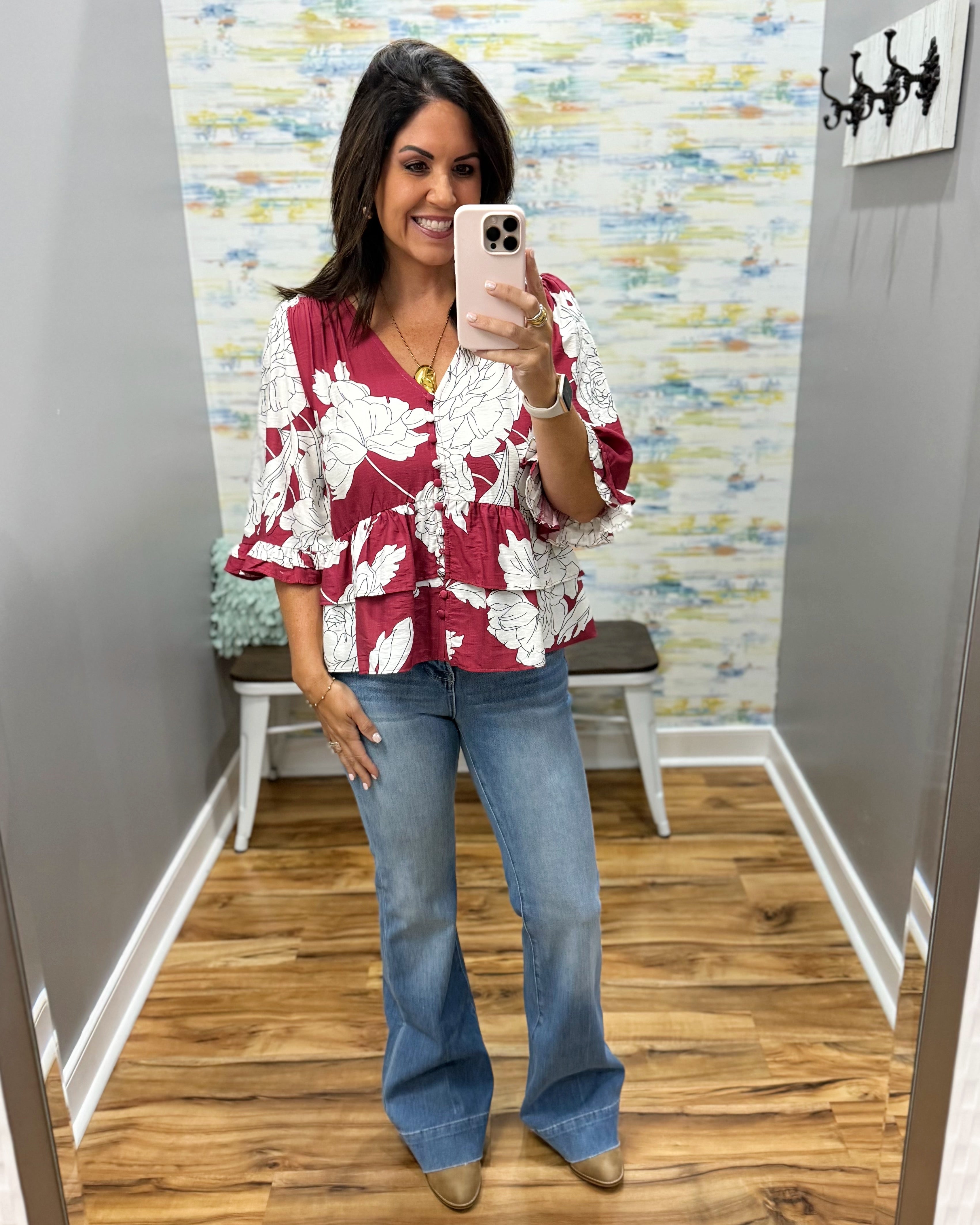 A woman takes a mirror selfie in a fitting room wearing a burgundy and white floral tiered blouse, blue flare jeans, and tan pointed-toe flats.