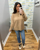 A woman takes a mirror selfie in a fitting room wearing a chunky knit crewneck sweater in a warm tan color, paired with blue jeans and black-and-white sneakers.