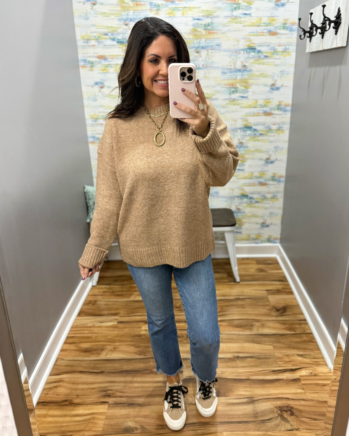 A woman takes a mirror selfie in a fitting room wearing a chunky knit crewneck sweater in a warm tan color, paired with blue jeans and black-and-white sneakers.