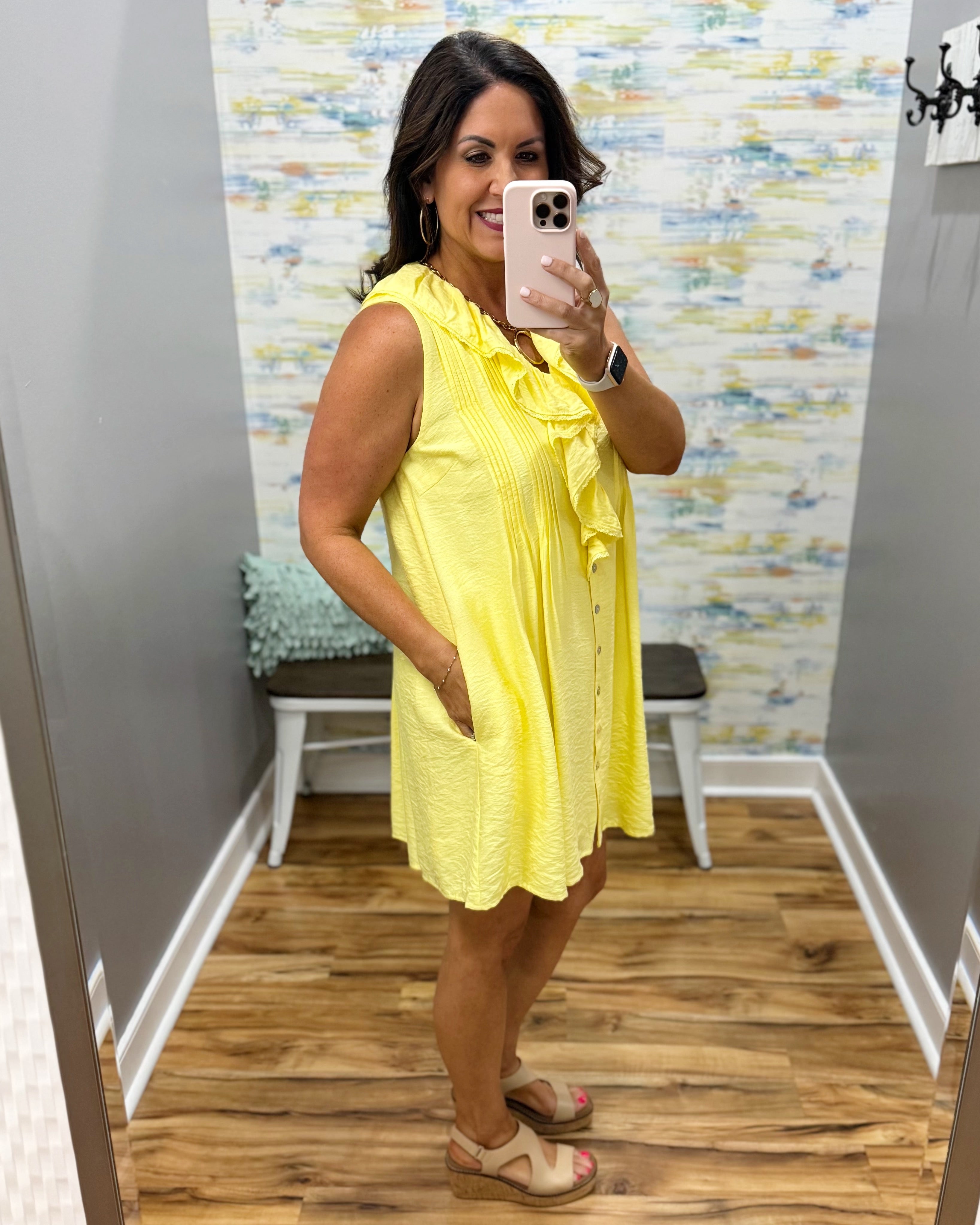 Woman wearing a yellow dress taking a mirror selfie in a room with a patterned wall.