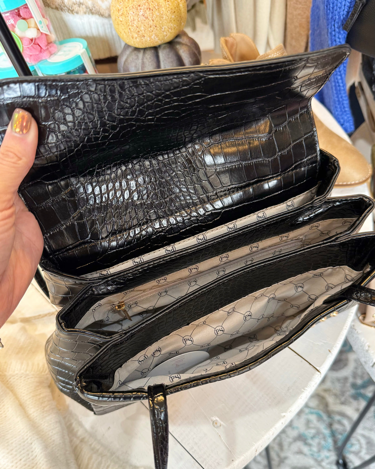 A close-up of a black crocodile-embossed leather handbag being held open by a hand, showing the interior lining with a repeating monogram pattern.