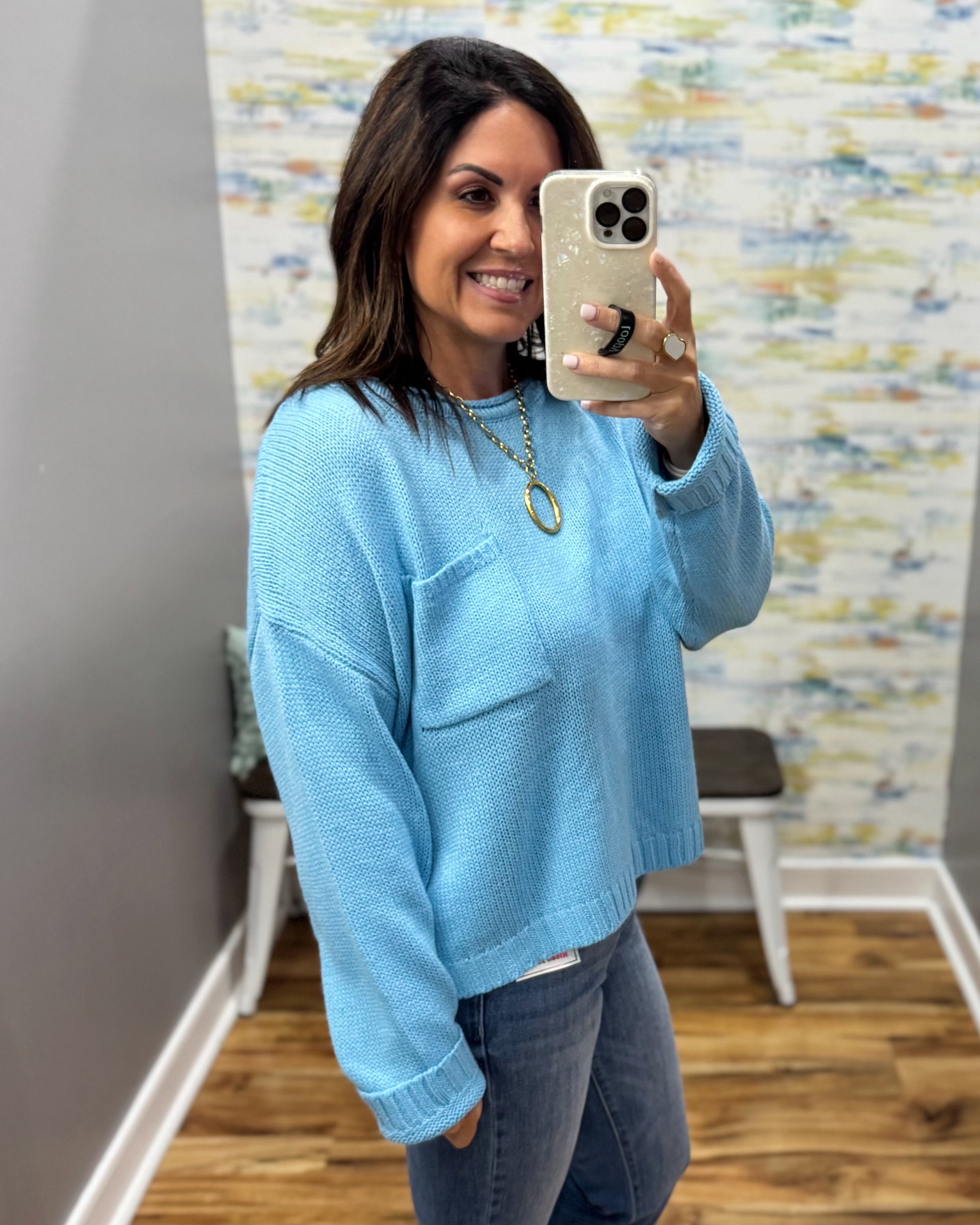 A woman takes a mirror selfie wearing a light blue knit sweater with a chest pocket and gray jeans in an indoor room with a patterned wallpaper background.