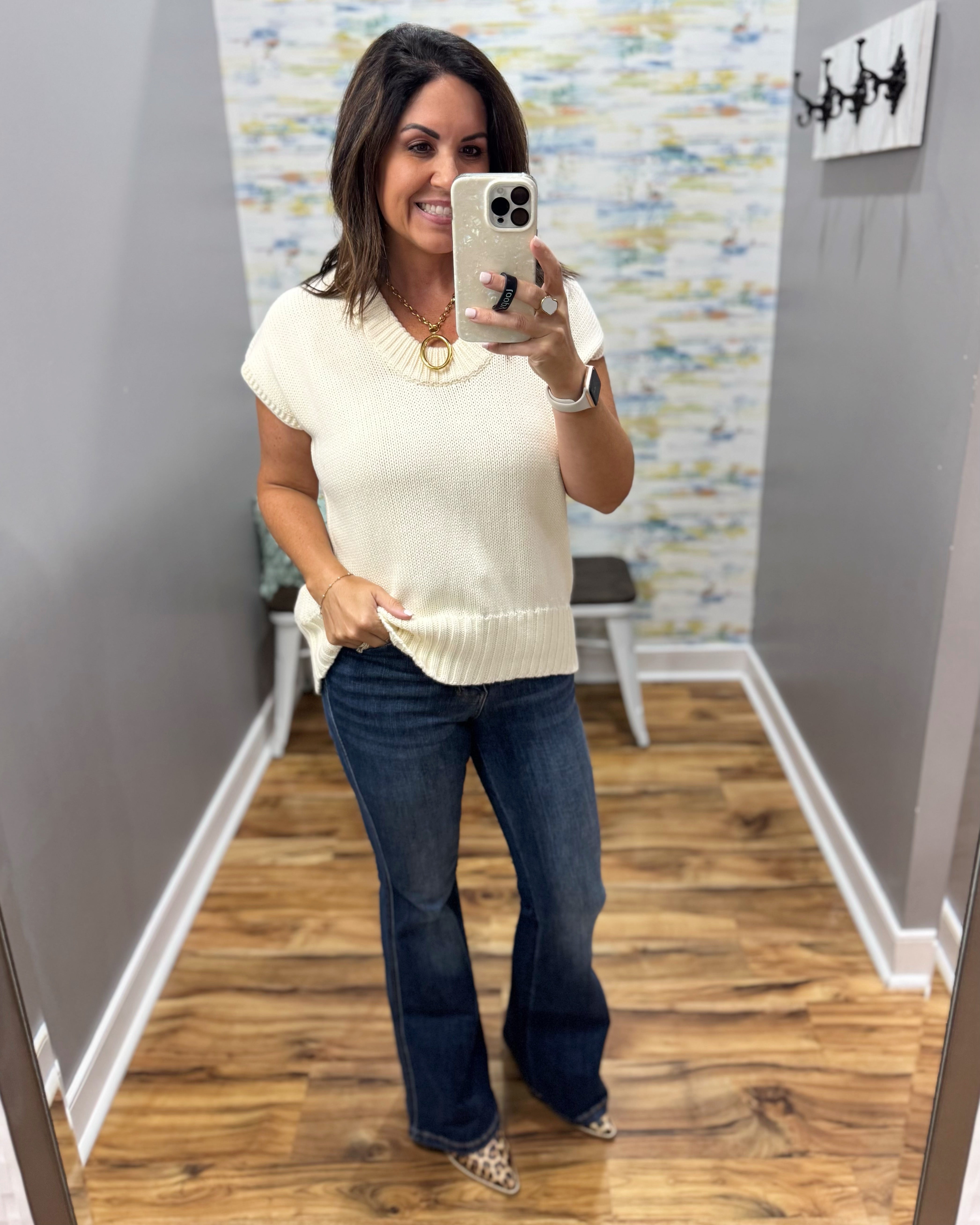 A woman takes a mirror selfie wearing a cream ribbed short-sleeve knit top and dark blue flare jeans in a fitting room with wood floors and gray walls.