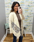 A woman takes a mirror selfie wearing a cream knit cardigan with a geometric patterned panel and fringe, layered over a brown top and dark jeans.