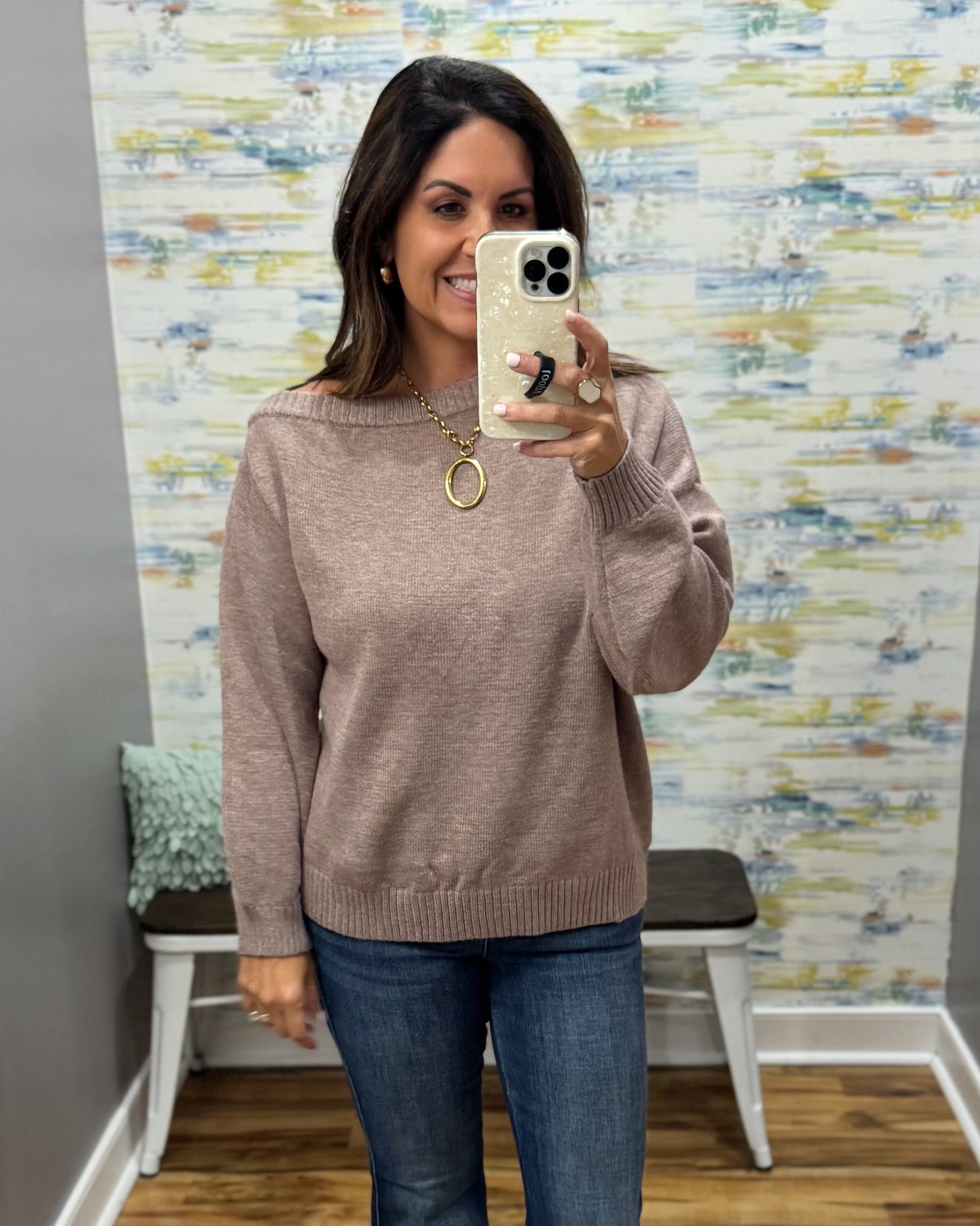 A woman takes a mirror selfie wearing a dusty rose knit sweater and blue jeans in front of a patterned wallpaper background.