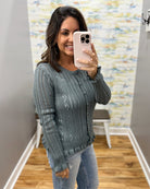 A woman takes a mirror selfie wearing a textured, ribbed knit cardigan sweater in a muted slate blue-gray color, paired with light-wash blue jeans.
