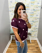 A woman takes a mirror selfie wearing a short-sleeve knit top in plum with pink and white polka dots and pink trim, paired with blue jeans.
