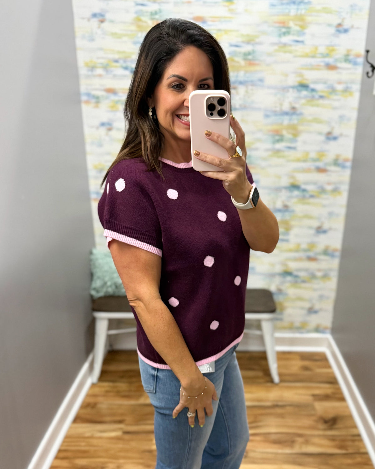 A woman takes a mirror selfie wearing a short-sleeve knit top in plum with pink and white polka dots and pink trim, paired with blue jeans.