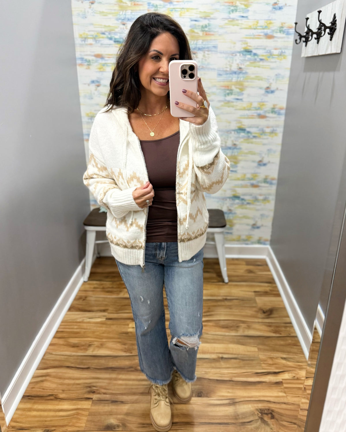 A woman takes a mirror selfie in a fitting room wearing a cream and tan patterned knit cardigan over a brown tank top, blue distressed jeans, and beige lace-up boots.