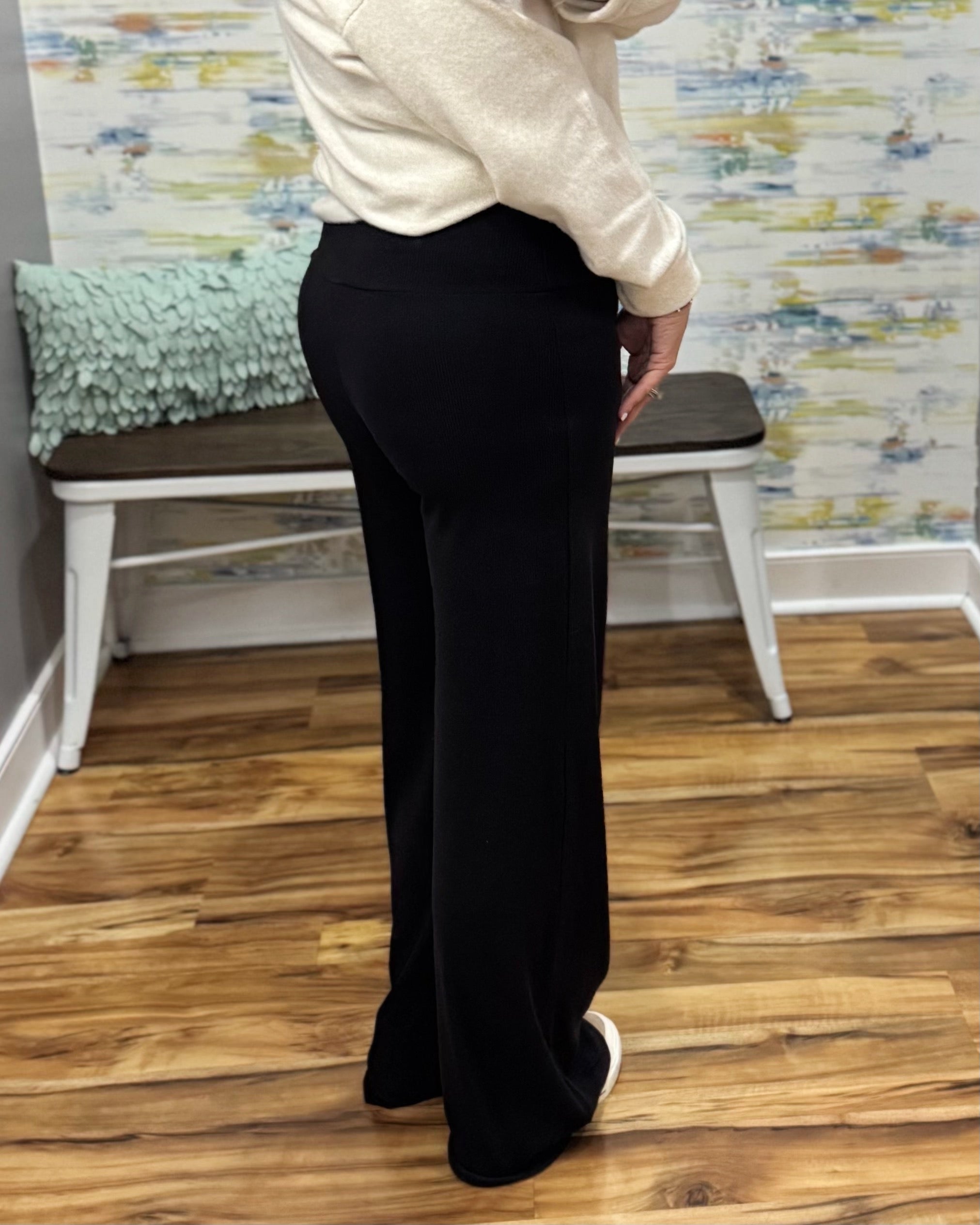 A person wearing black wide-leg pants and a cream sweater, standing in a room with wood floors and a bench in the background.