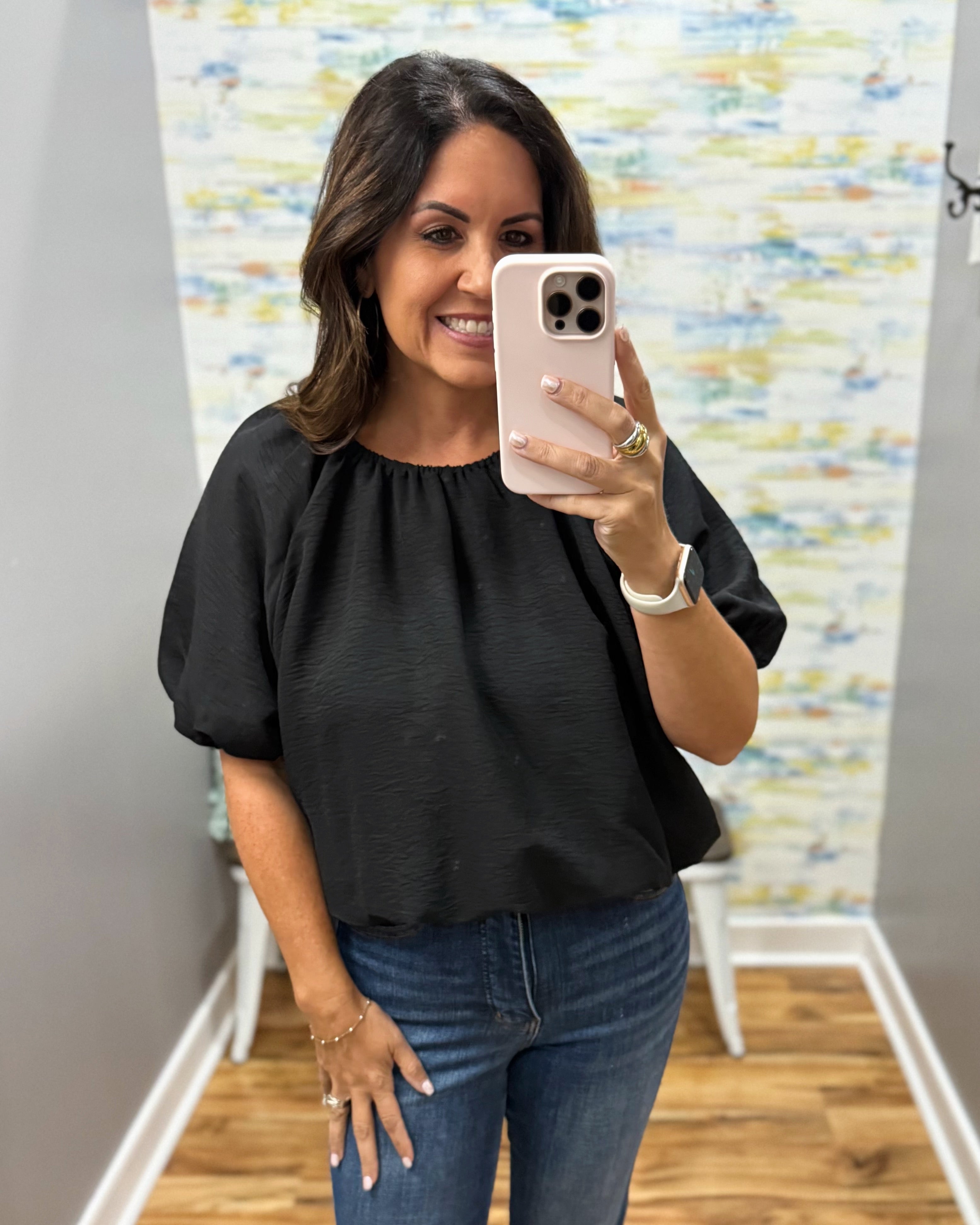 A woman takes a mirror selfie wearing a black short-sleeve top with a gathered neckline and blue jeans, holding a light pink smartphone in front of her face.