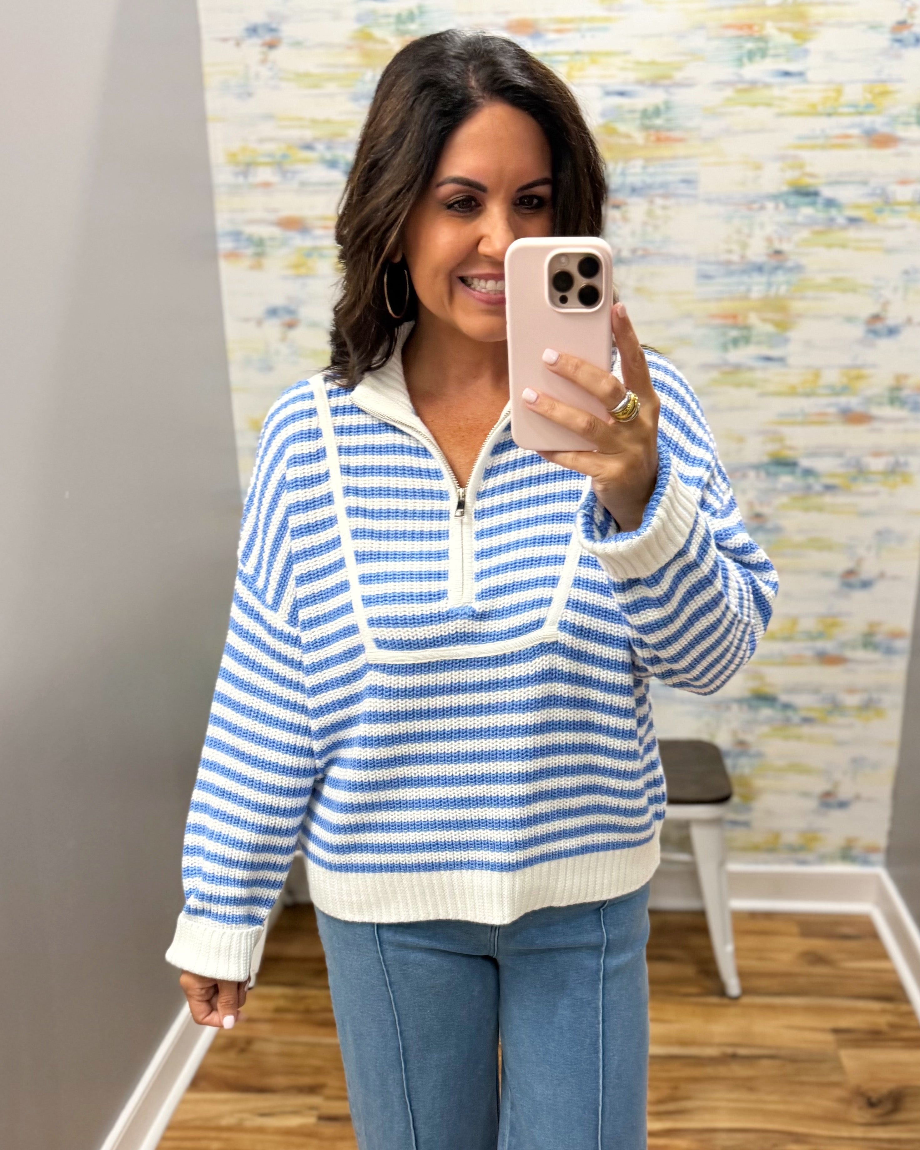 A woman takes a mirror selfie wearing a blue and white striped knit half-zip sweater and blue jeans in an indoor room with a patterned wall and wood floor.