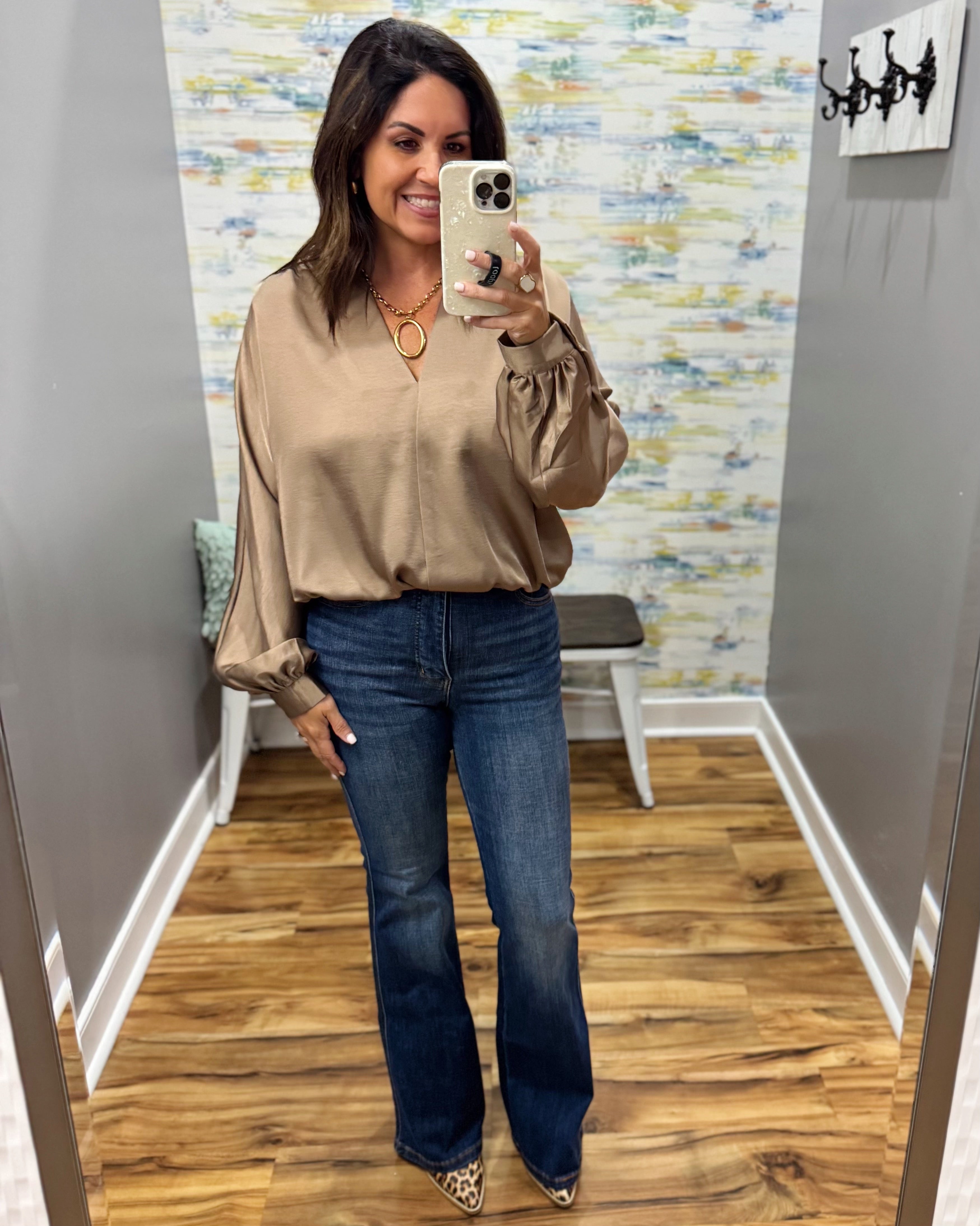 A woman takes a mirror selfie in a fitting room wearing a beige satin blouse with puff sleeves and dark blue flare jeans.