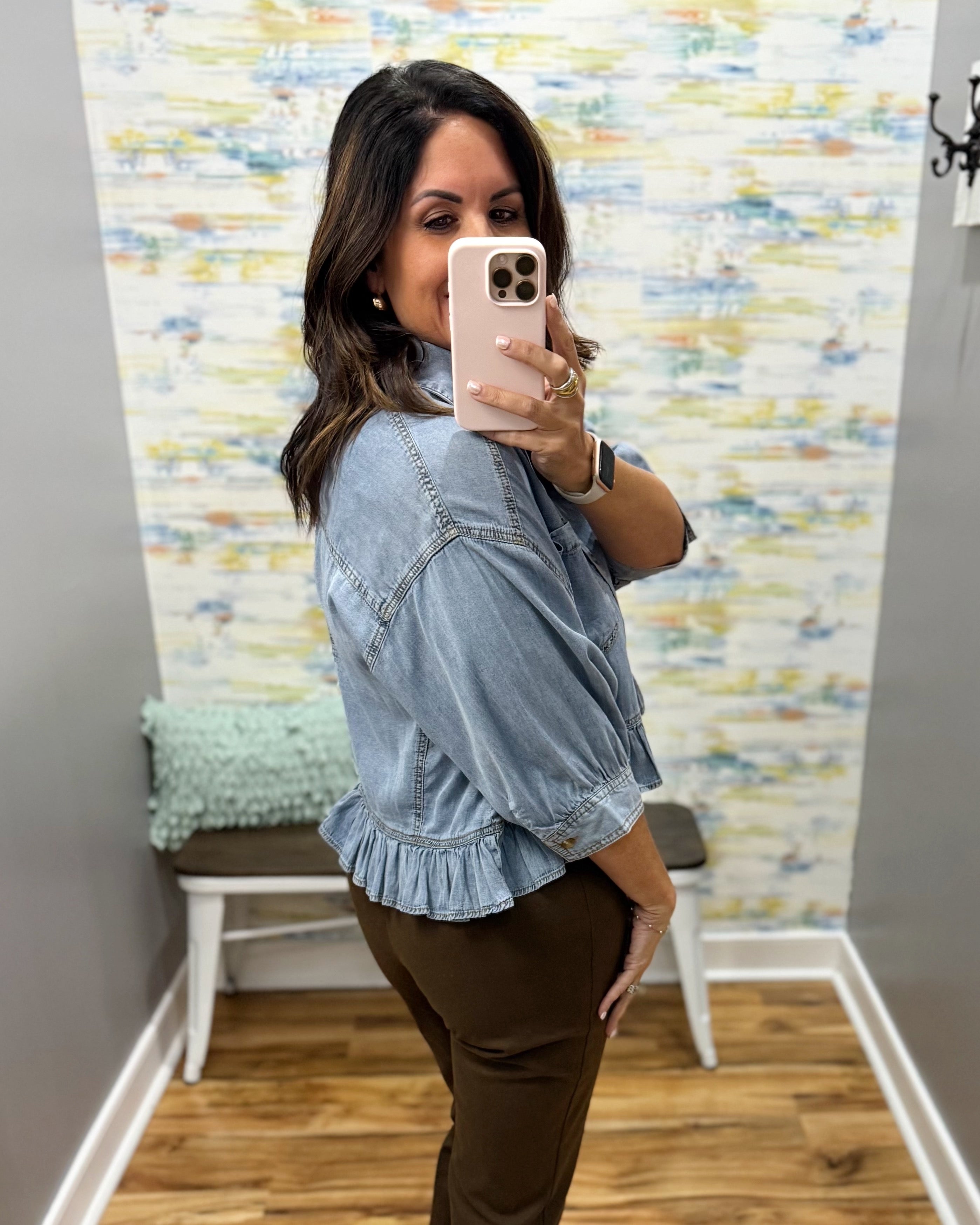 A woman takes a mirror selfie wearing a light blue denim ruffle-sleeve blouse and brown pants in a home setting with a patterned wallpaper background.