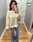 A woman takes a mirror selfie wearing a cream and tan striped knit sweater, blue jeans, and brown boots in a home setting with wood floors and gray walls.