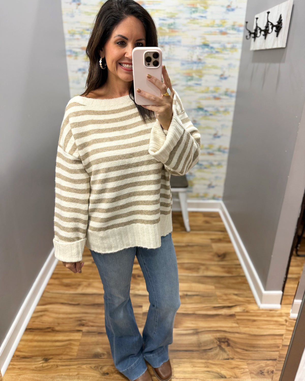 A woman takes a mirror selfie wearing a cream and tan striped knit sweater, blue jeans, and brown boots in a home setting with wood floors and gray walls.