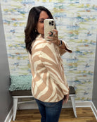 A woman takes a mirror selfie wearing a beige and cream zebra-patterned cardigan sweater, paired with blue jeans, in front of a patterned wallpapered wall.