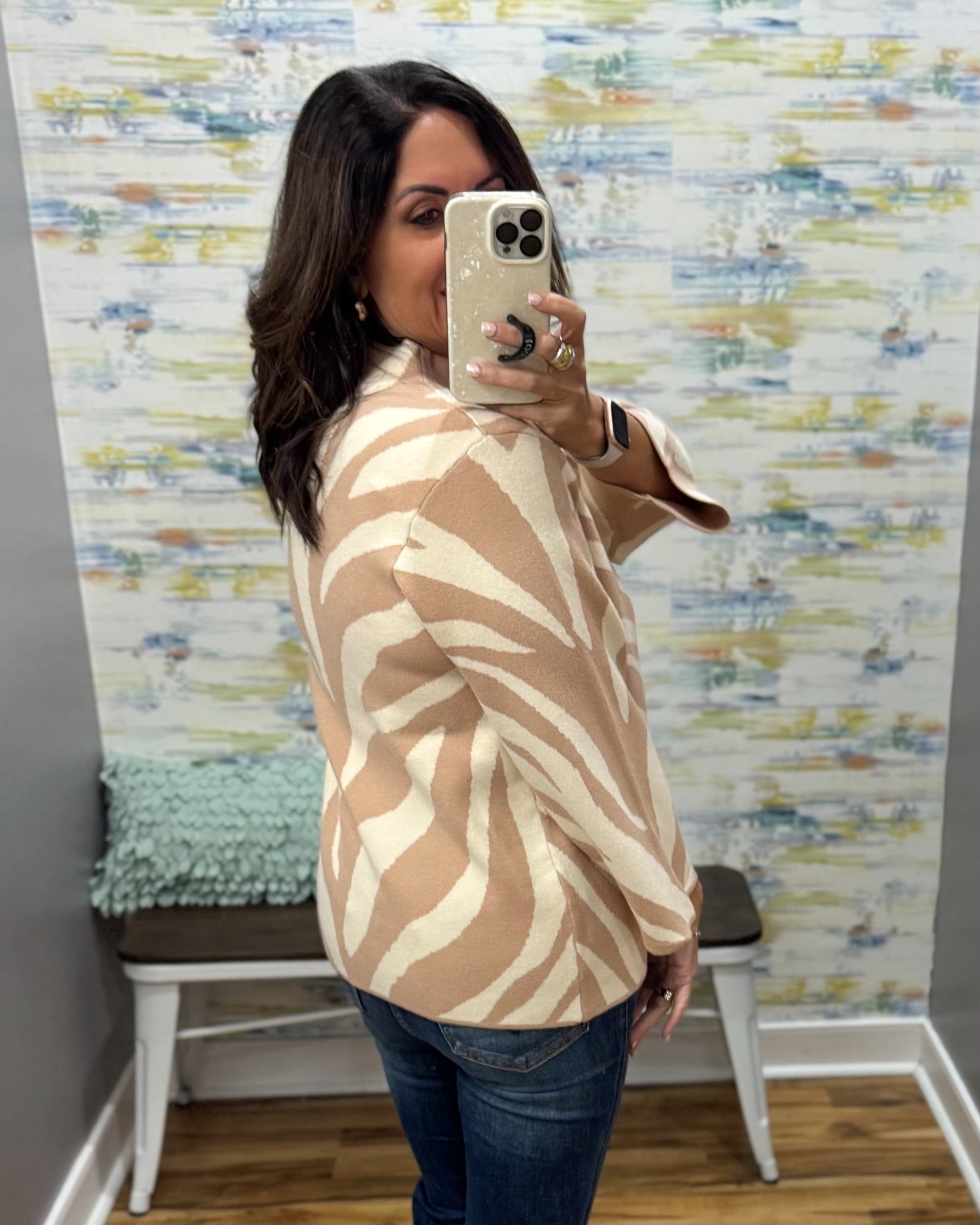 A woman takes a mirror selfie wearing a beige and cream zebra-patterned cardigan sweater, paired with blue jeans, in front of a patterned wallpapered wall.