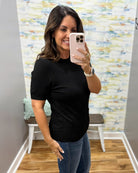 A woman taking a mirror selfie in a fitting room wearing a black short-sleeve crewneck t-shirt and blue jeans.