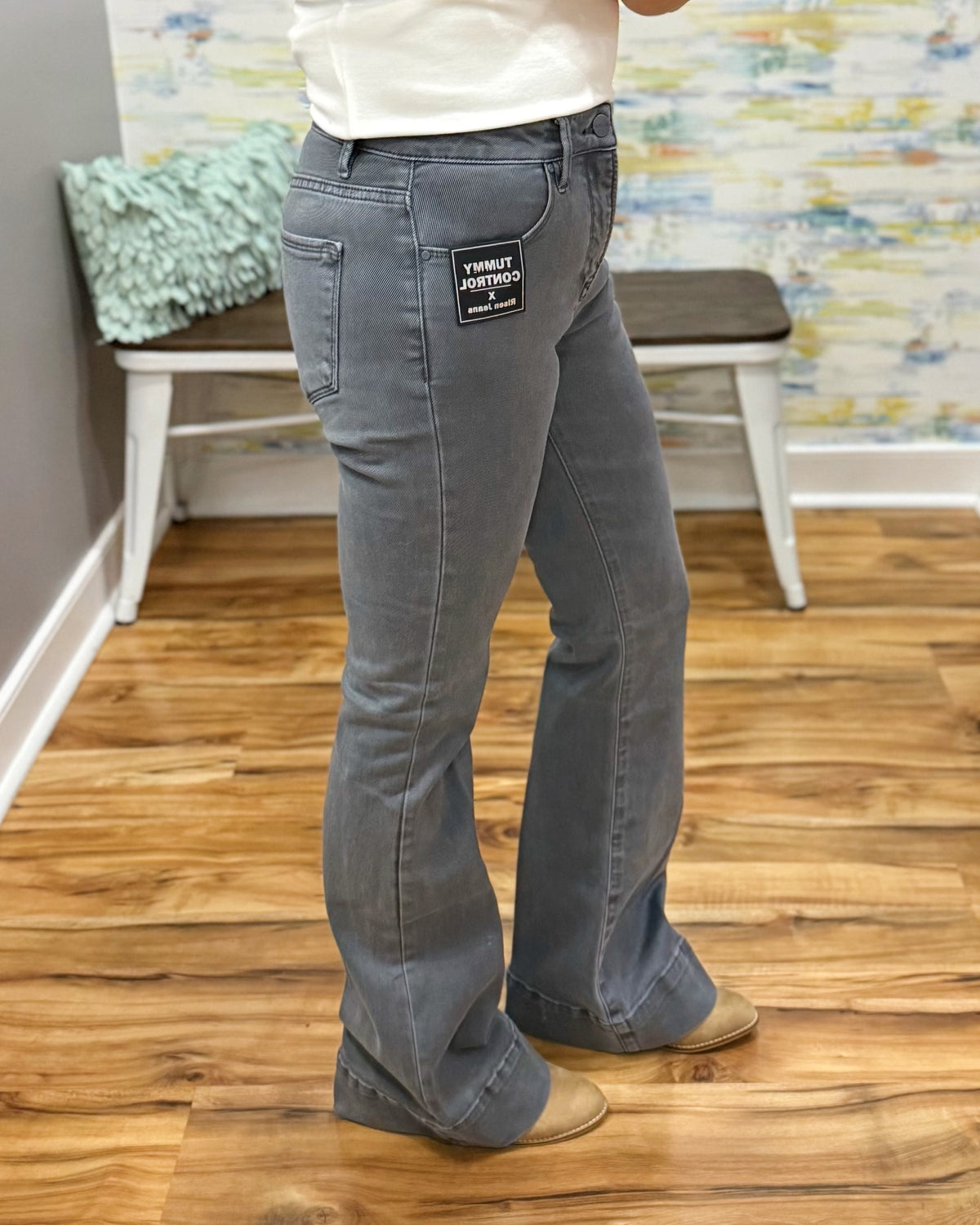 A person wearing faded medium-wash blue flare jeans with a black 'SWEATWEAR JERSEY' patch on the back pocket, standing on a wooden floor in an indoor room.