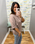 A woman takes a mirror selfie wearing a loose, short-sleeve knit sweater in a dusty mauve-pink color and light-wash blue jeans in a home entryway.