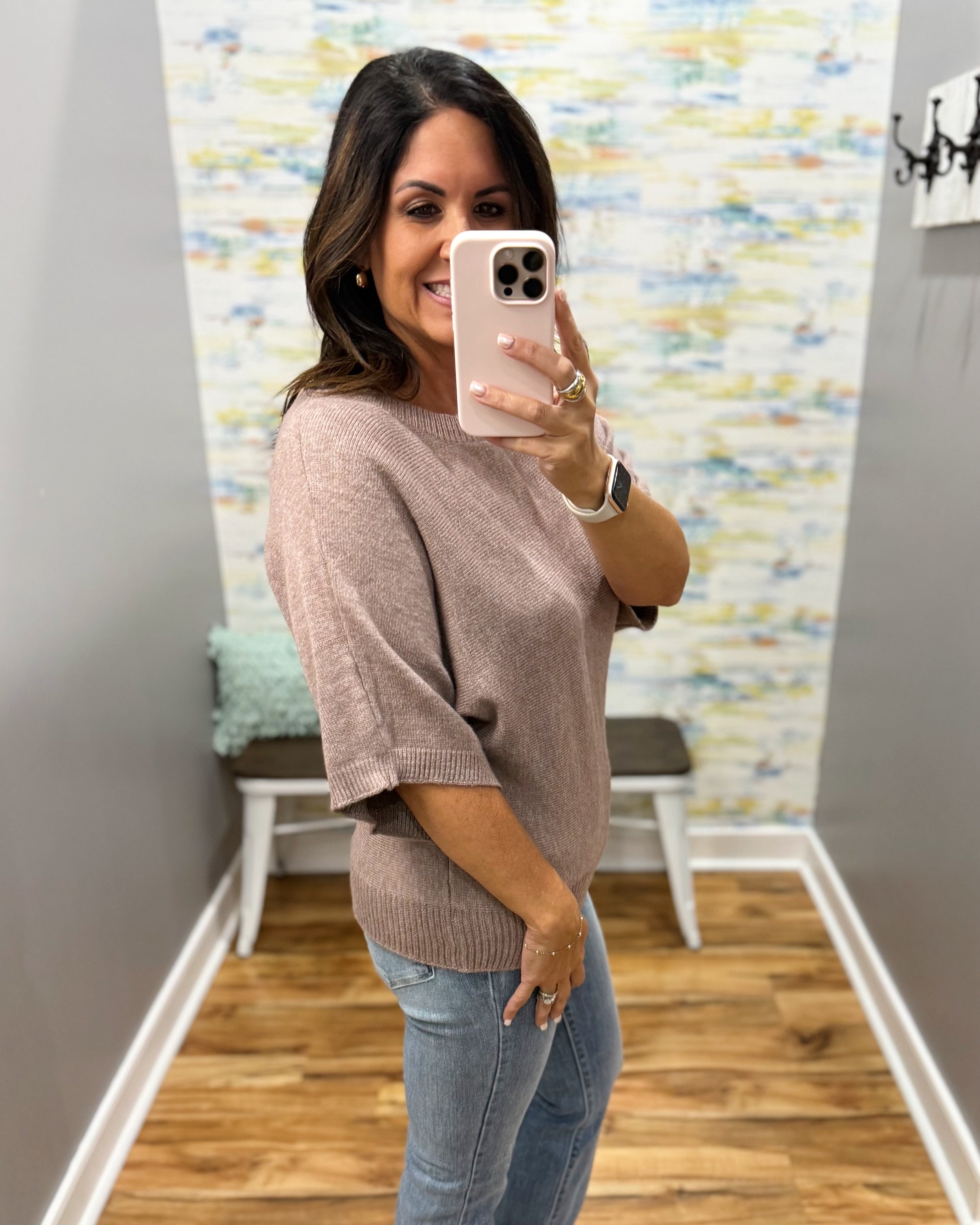 A woman takes a mirror selfie wearing a loose, short-sleeve knit sweater in a dusty mauve-pink color and light-wash blue jeans in a home entryway.