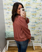 A woman takes a mirror selfie wearing a dusty rose satin bomber jacket and blue jeans in front of a patterned wallpaper background.