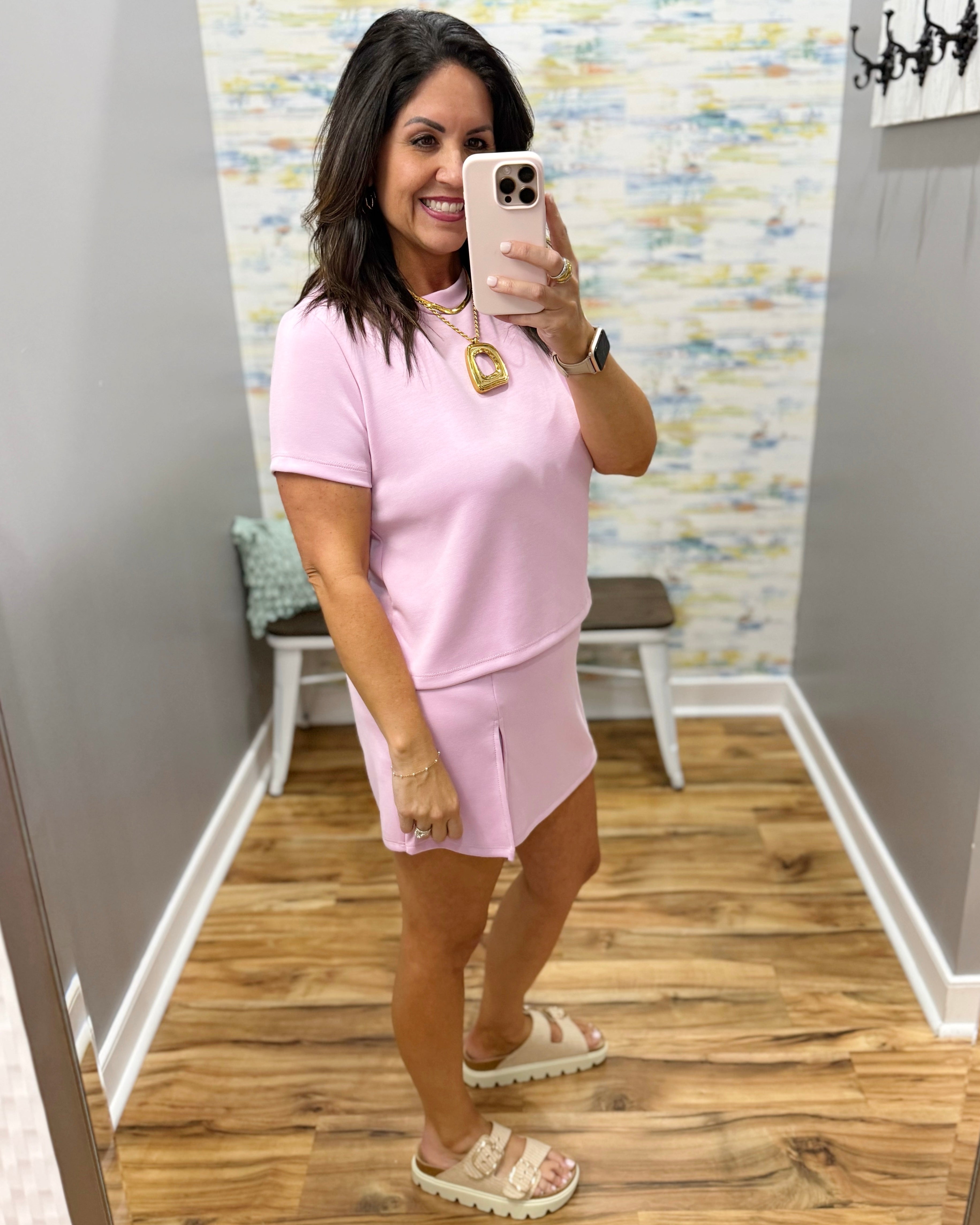 Woman taking a mirror selfie wearing a pink outfit in a room with a patterned wall.