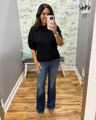 A woman takes a mirror selfie showing an outfit consisting of a black short-sleeve mock neck top and blue flare jeans, standing in a fitting room with wood floors and patterned wallpaper.