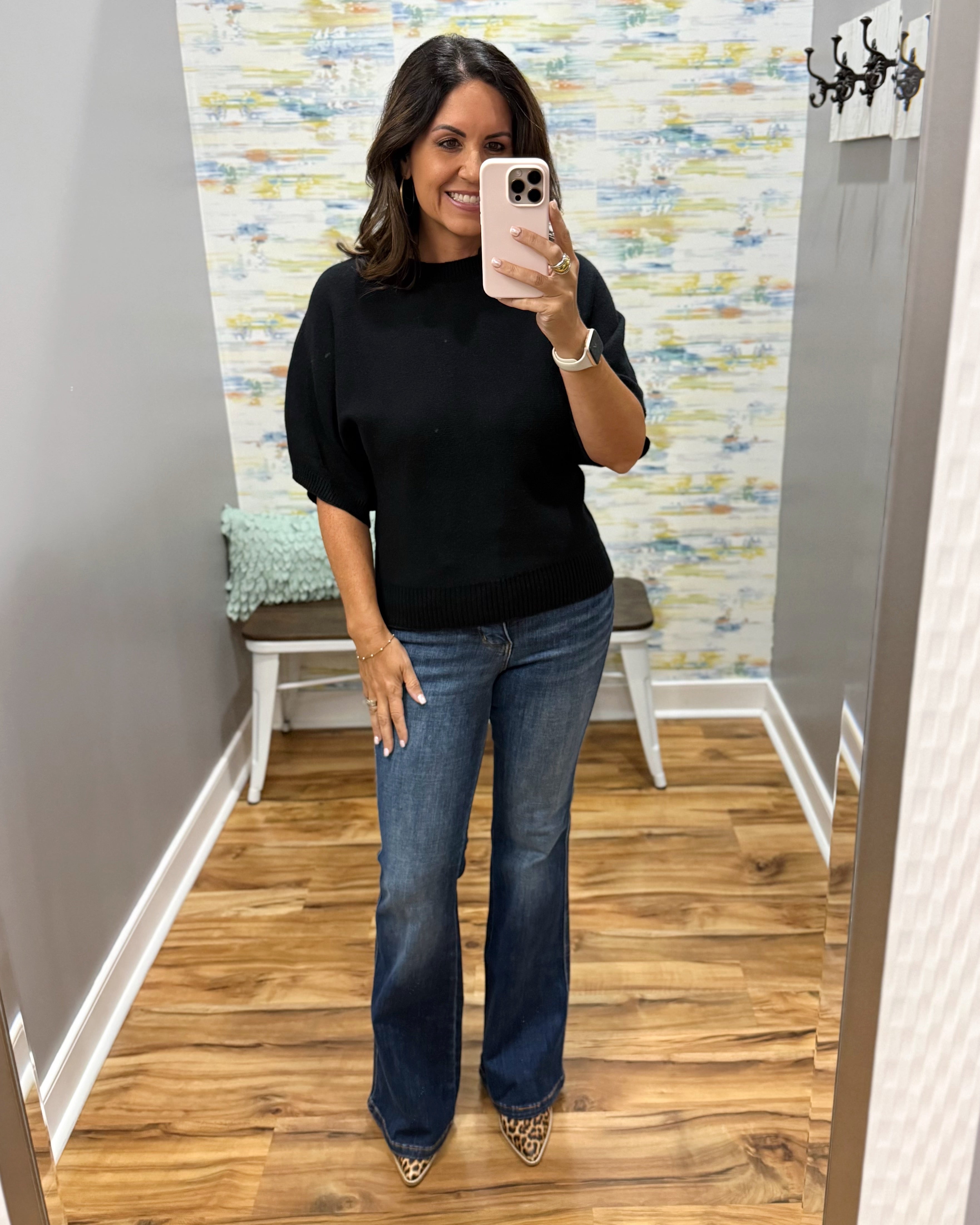 A woman takes a mirror selfie showing an outfit consisting of a black short-sleeve mock neck top and blue flare jeans, standing in a fitting room with wood floors and patterned wallpaper.