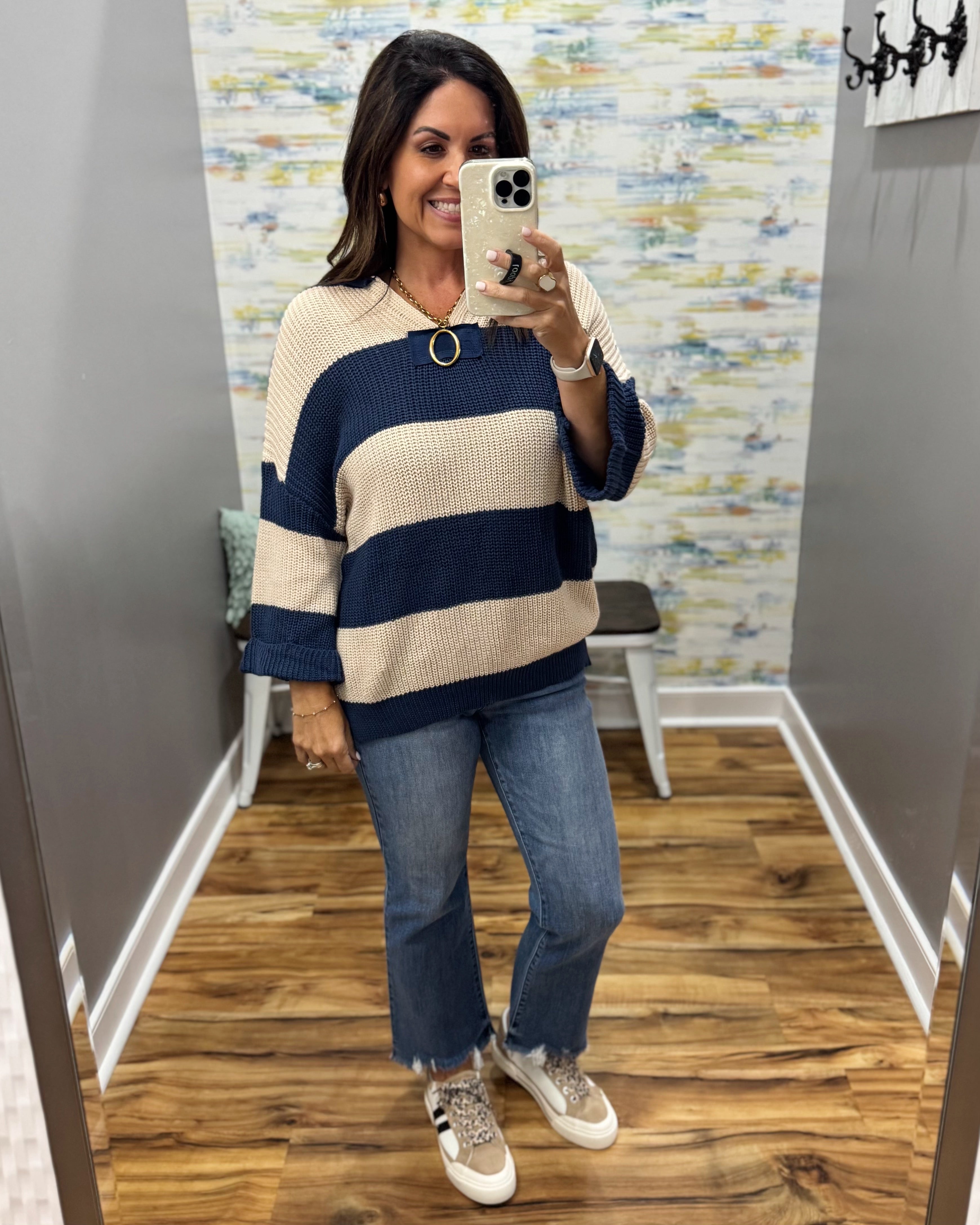 A woman takes a mirror selfie in a fitting room wearing a waffle-knit sweater with horizontal navy and cream stripes, blue flare jeans, and beige sneakers.