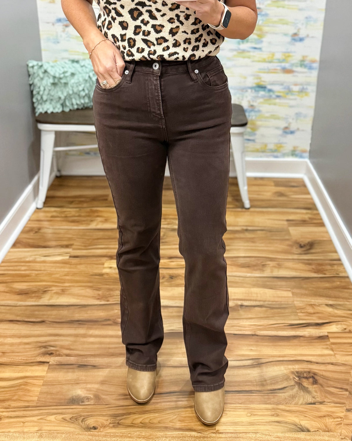 A person wearing mica dark brown straight-leg jeans, a leopard-print top, and tan boots, standing on a wood floor in an indoor room.