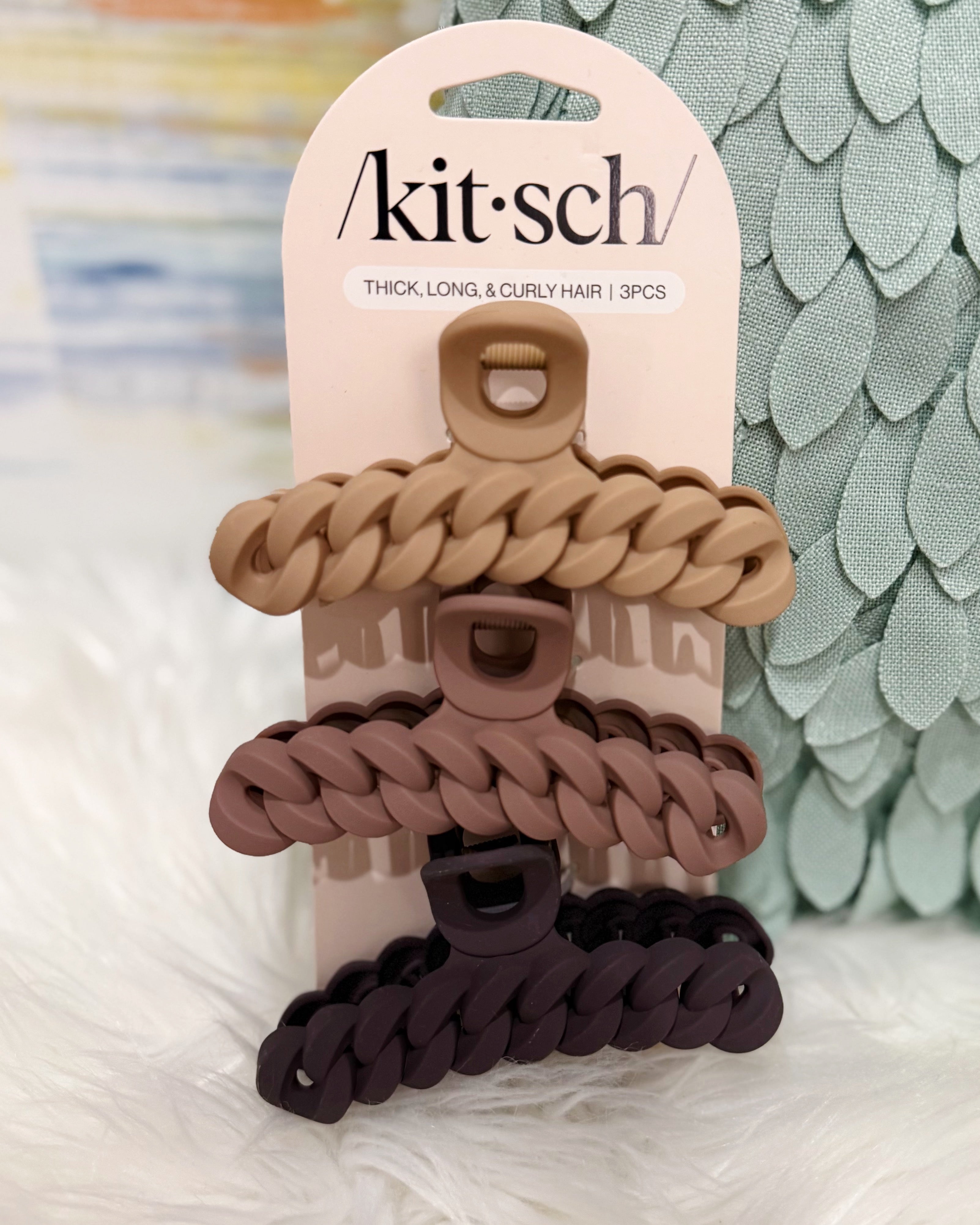 A 3-piece hair claw clip set by kit·sch on its original cream-colored cardboard hanger. The set includes three thick, braided claw clips in light tan, dusty rose, and dark plum purple, displayed against a soft white faux fur background with a green scale-patterned box in the background.