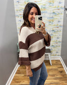 A woman takes a mirror selfie wearing a brown and cream striped knit sweater with elbow-length sleeves and blue jeans in an indoor room with wood floors and a patterned wall.