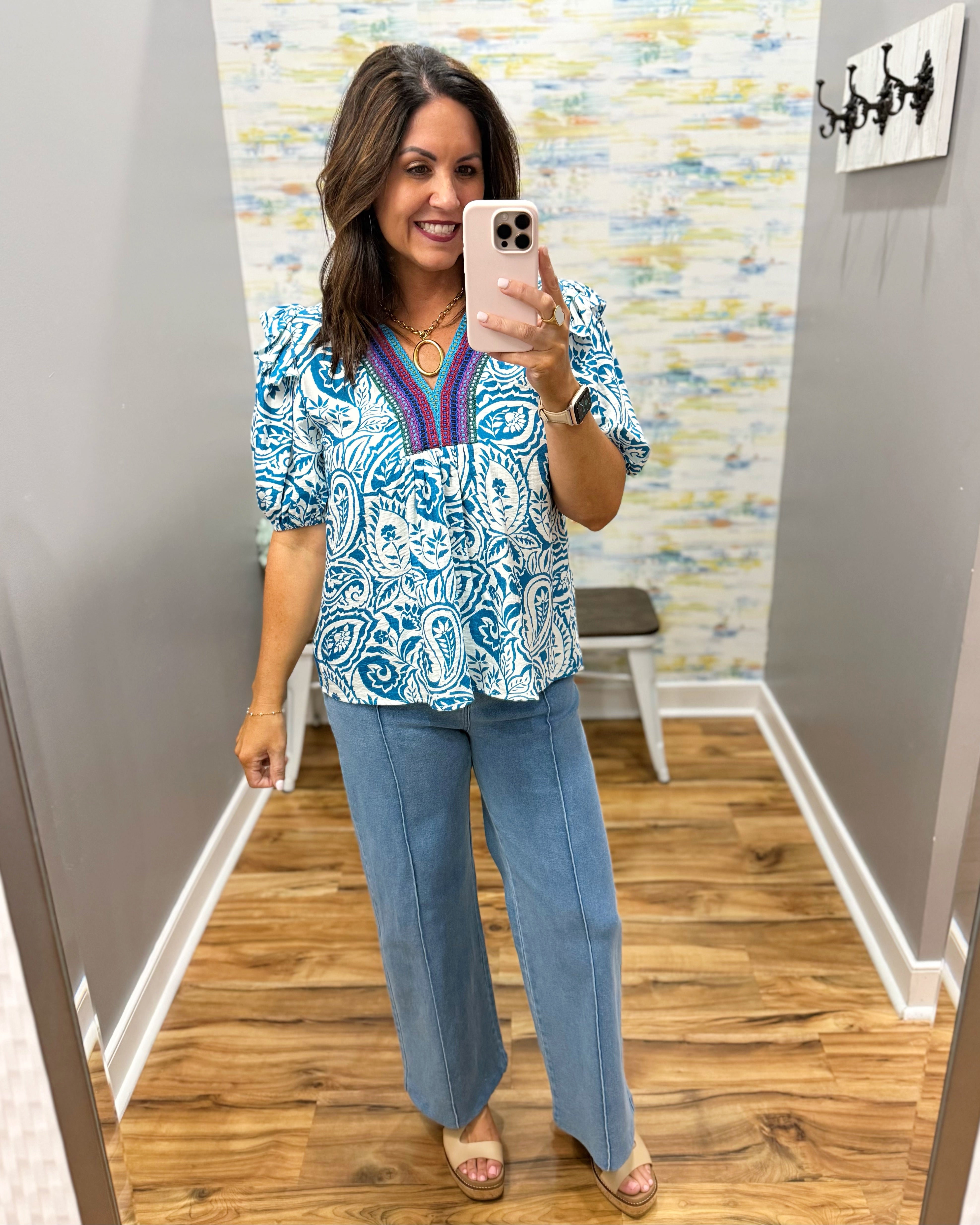 Woman taking a mirror selfie wearing a blue and white patterned top and light blue jeans in a room with a colorful wall.