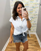 Woman taking a mirror selfie wearing a white striped shirt and denim shorts in a room with a colorful wall.