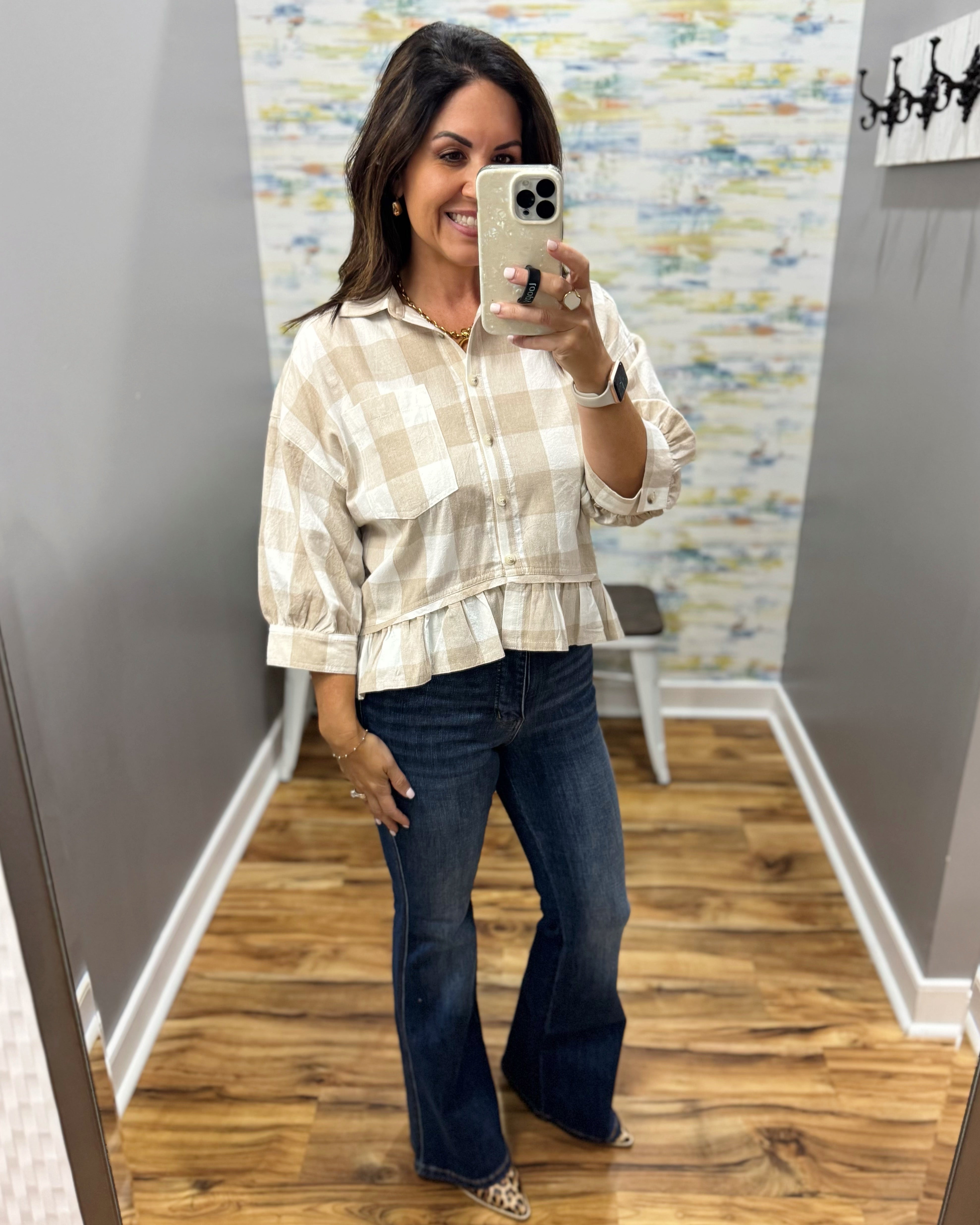 A woman takes a mirror selfie wearing a beige and white gingham check long-sleeve blouse with a ruffled hem and dark blue flare jeans in a fitting room.